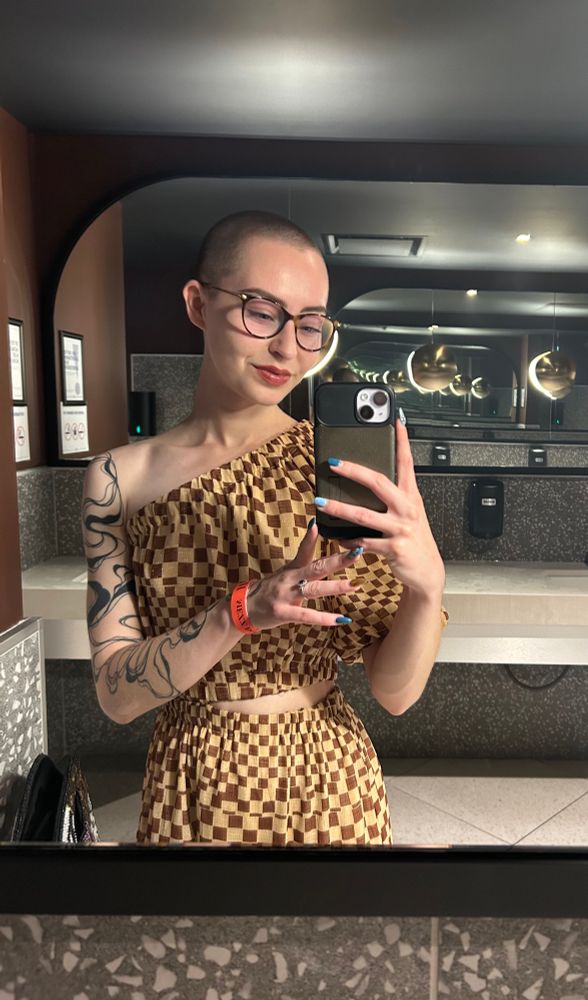 Darla stands in a pristine washroom at an event and takes a selfie in the mirror. She is wearing a checkered maxi dress with only one sleeve and a slit at the waist on the opposite side. She is smiling contentedly. 