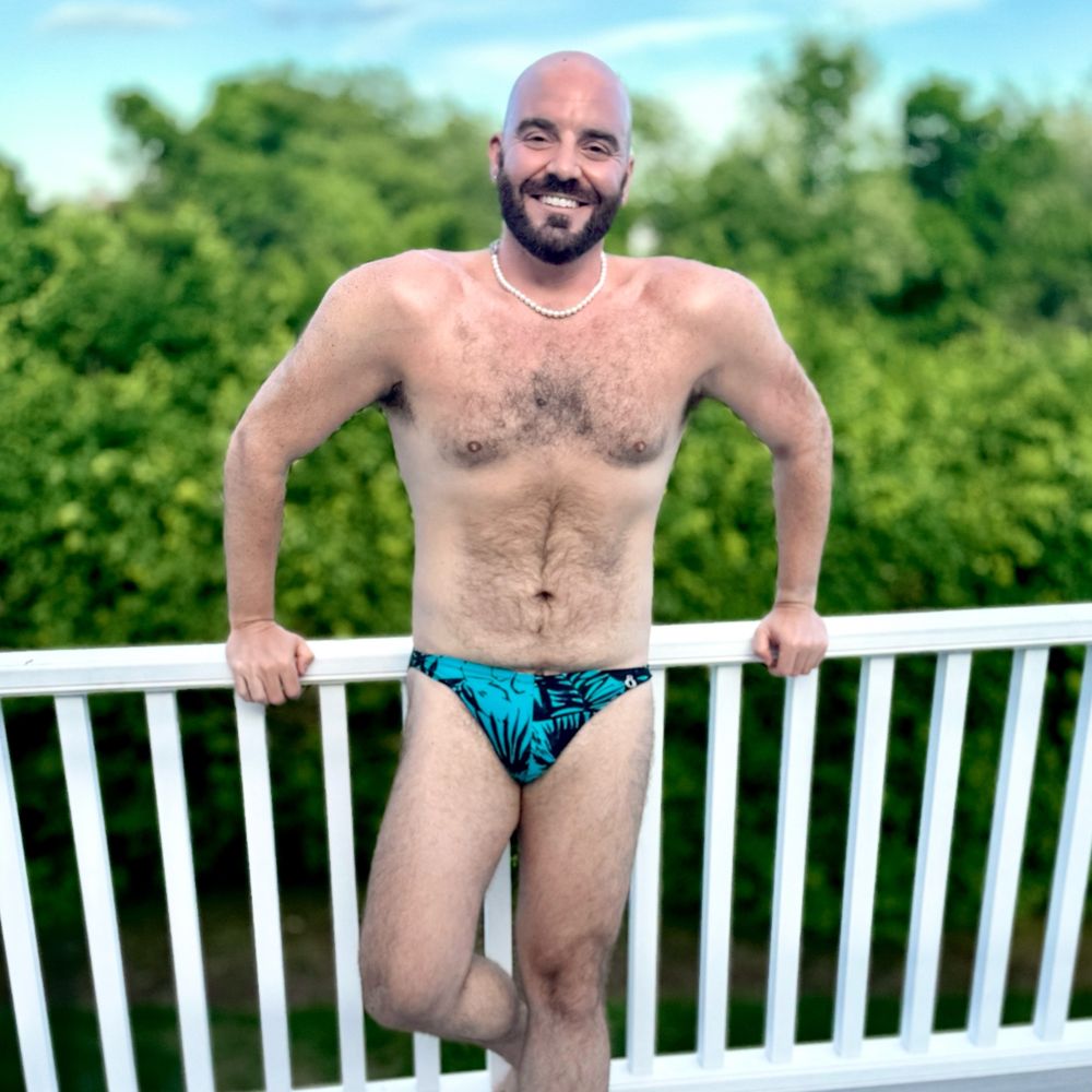 Me, wearing swim briefs and a pearl necklace, posing on a deck