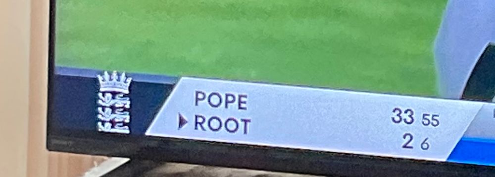 A cropped photo of a tv, showing only the bottom left of the screen. It shows the current England cricket batters’ surnames: Pope and Root