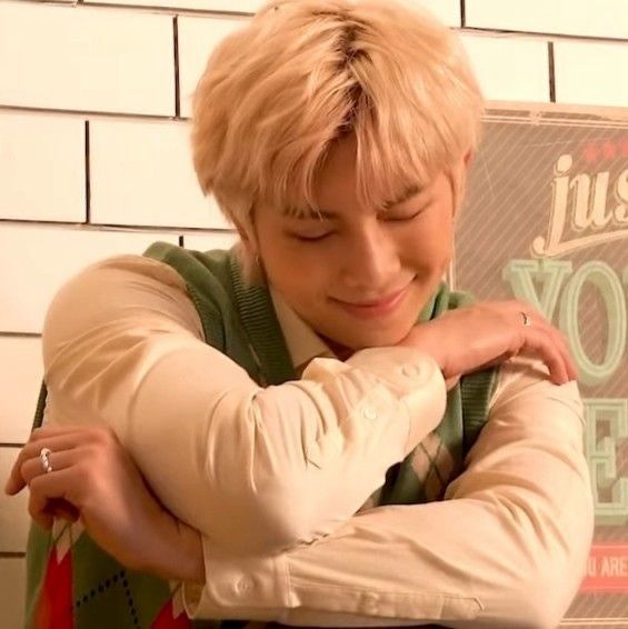 a screenshot of namjoon during a photoshoot, his hair is blonde, he’s wearing a white dress shirt with a green, red, and white vest over top of it. he has a silver hoop in his ear and silver rings on each hand. his arms are crossed at an angle, his chin leaned down so it’s laying against one of his wrists, his eyes closed, smiling, the cutest dimple on display 
