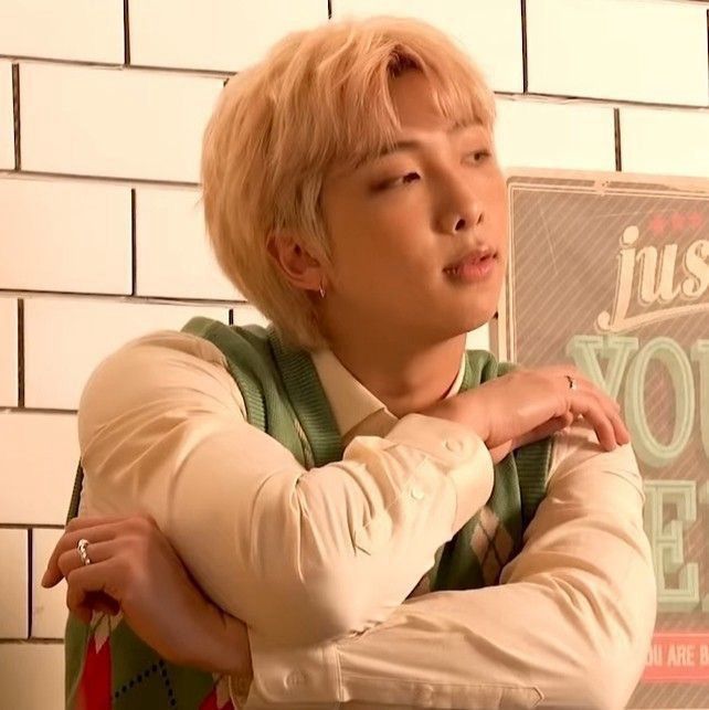 a screenshot of namjoon during a photoshoot, his hair is blonde, he’s wearing a white dress shirt with a green, red, and white vest over top of it. he has a silver hoop in his ear and silver rings on each hand. his arms are crossed at an angle, his head slightly tilted as he looks off to the right 