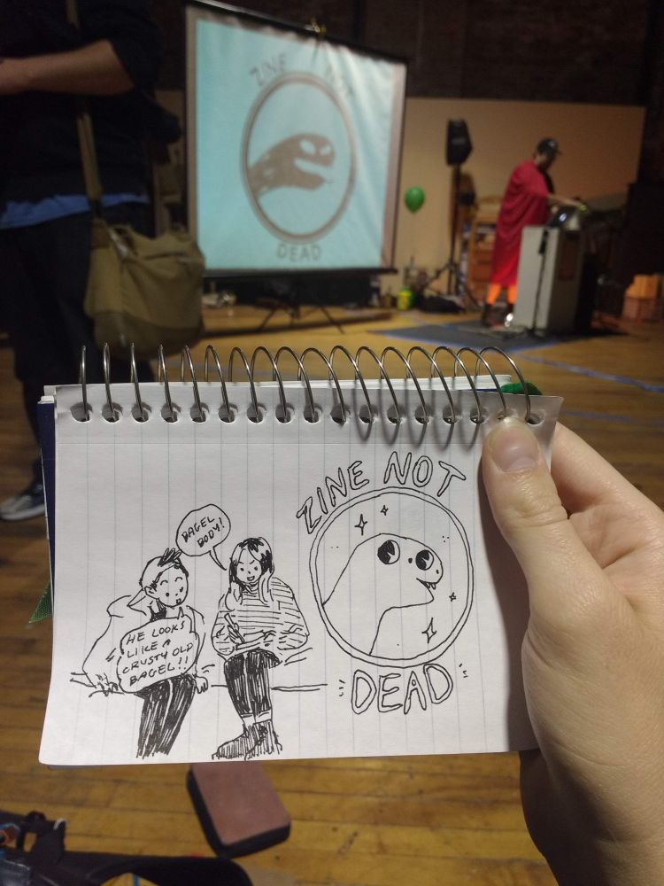 I am holding a small spiral bound- lined notebook, the drawing on it depicts two friends drawn in pen reacting to a drawing they are doing with the zine not dead logo reamigined next to it.