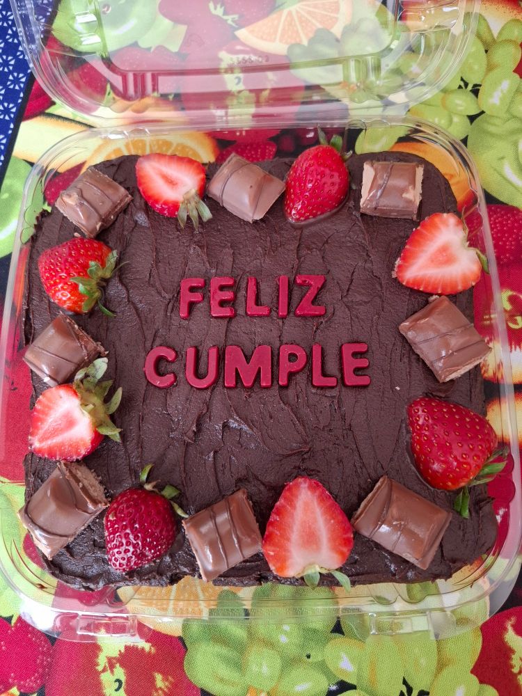 Brownies covered in chocolate frosting and decorated with strawberry halves and kinder bueno chocolate pieces. "Feliz cumple" is written in the middle with red colored chocolate letters. Note: "feliz cumple" is a shortened version of "Happy birthday" in Spanish. 