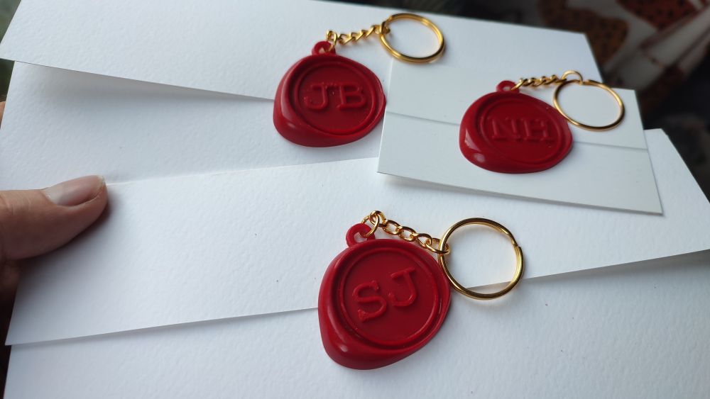 Three wax seal style keyrings with different pairs of initials, two sealing a folded taskmaster task and one on a business card printed like a task