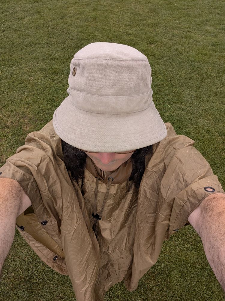 A man in a soaked nylon rain poncho and a Tilley hat holds his bare arm up to support the camera for a selfie. His face is hidden, but you can see his long brown hair and thick glasses