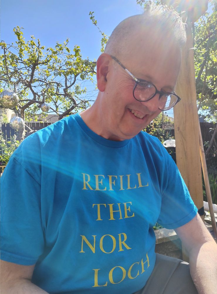 Man (my man!) wears blue t-shirt with yellow lettering reading REFILL THE NOR LOCH (an ancient body of water in Edinburgh where Princes Street Gardens is now).