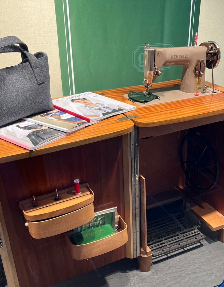 Eine alte Tretnähmaschine der Firma Singer. Links im Bild die Nähmaschine, rechts davon ein paar Modehefte und eine graue Tasche. Im Unterbau der Nähmaschine sieht man die Tretvorrichtung, links davon aufgesetzte Fächer für Nähgarn und kleine Nähutensilien. 