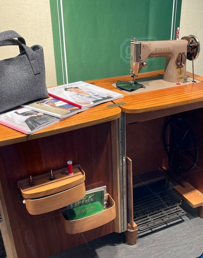 Eine alte Nähmaschine mit geöffnetem Unterbau. Im linken Teil kleine Ablageboxen für Nähutensilien. Auf dem oberen Nähtisch liegen Modezeitschriften und eine graue Stofftasche.  