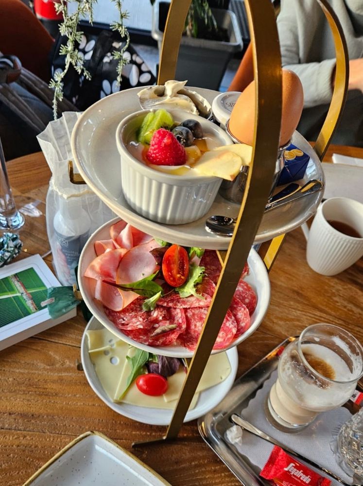 Auf einem Holztisch steht eine Etagere mit verschiedenen Lebensmitteln wie Käse, Schinken. Wurst, einem Ei und Früchten. Weiters auf dem Tisch kleine Tabletts mit Kaffeetassen und Gläser. 