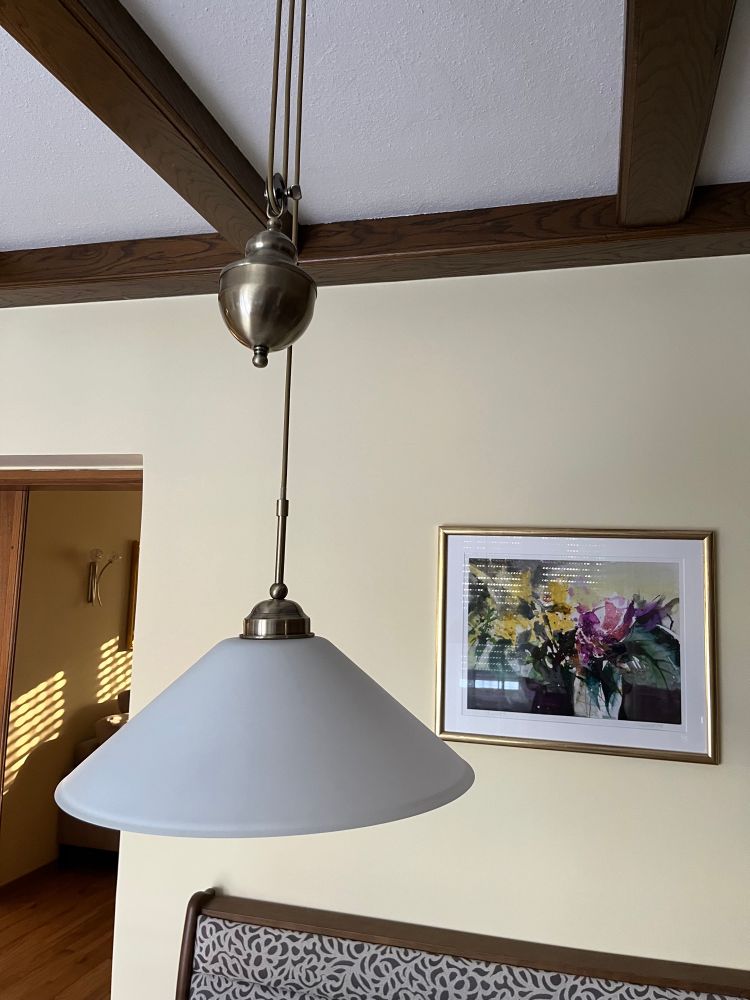 Eine weiße rustikale Pendellampe die je nach Bedarf hoch und runter gezogen werden kann. An der Decke dunkelbraune Holzbalken, dahinter eine gelb gestrichene Wand mit einem Bild in einem Goldrahmen. 