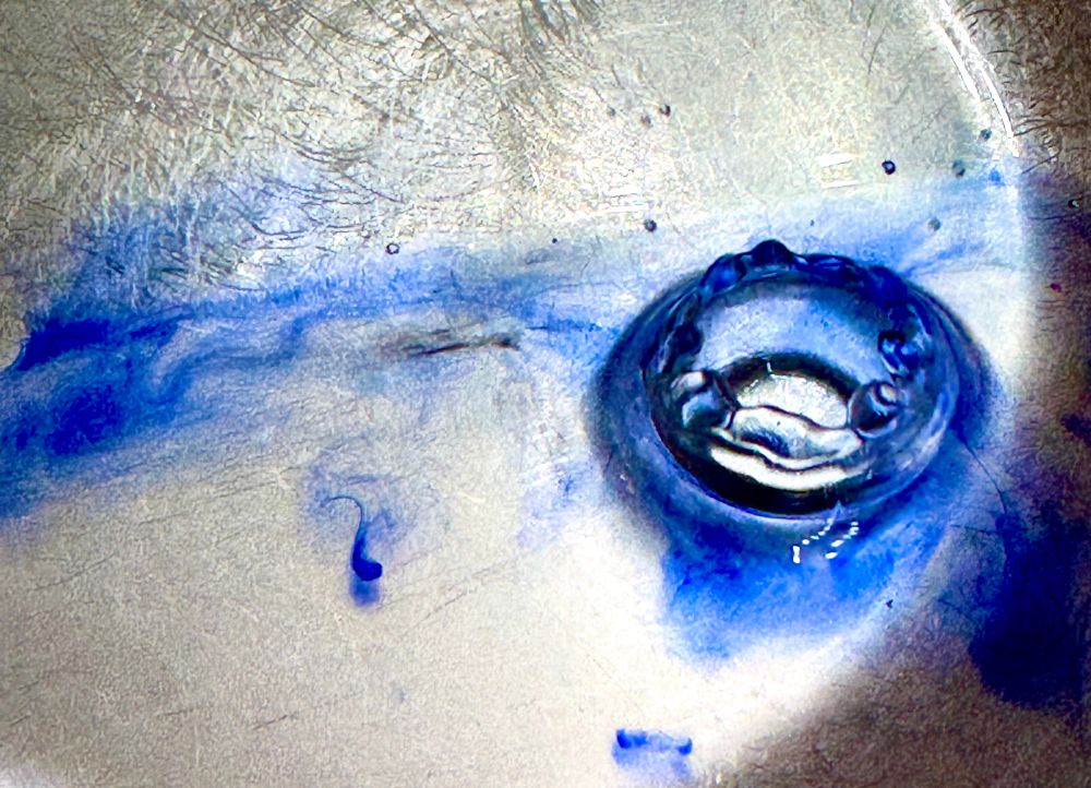 A drop of water impacts a pool of water in a stainless steel receptacle. The impact causing a smooth irruption of water. Deep blue ink is randomly scattered about the scene like whispy clouds. A half circle moon-like streak of white light frames the right side just near the impact site. Right of the semi circle the image fades to black with little water droplets attached to the container. The impact itself is awash in blue tones from deep to light to black and resembles an impact crater on the surface of the moon. 