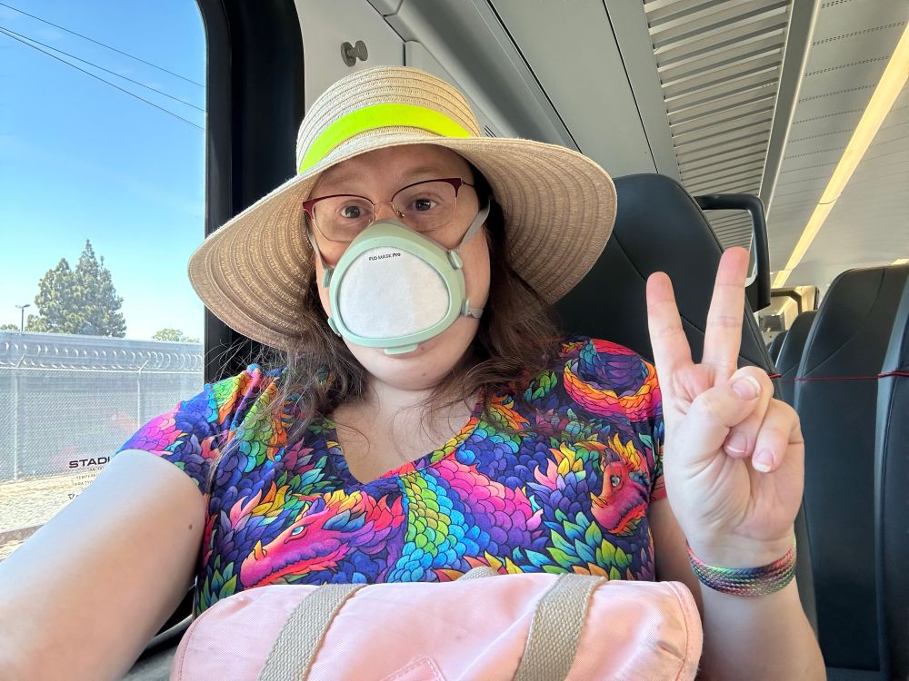 Liz in a straw sunhat, flo mask, dress wirh rainbow scaled dragons it riding on a train and waving a peace sign.