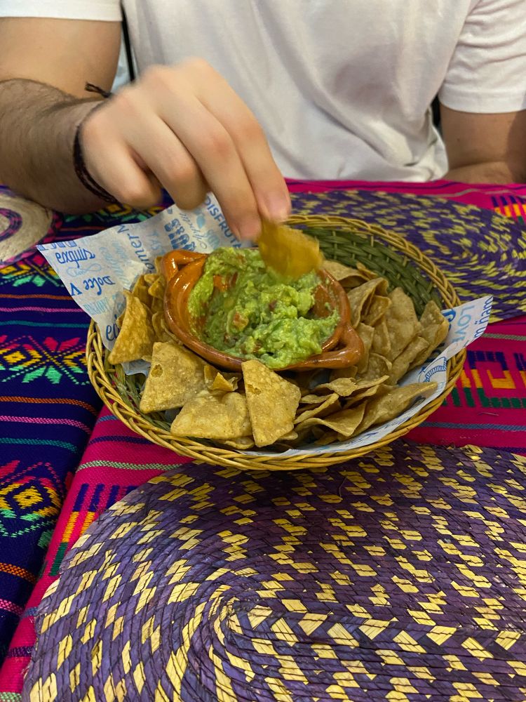 On a colourful ta me, a bowl of fried nachos and inside it, another smaller bowl with guacamole. A hand is seen picking up a nacho