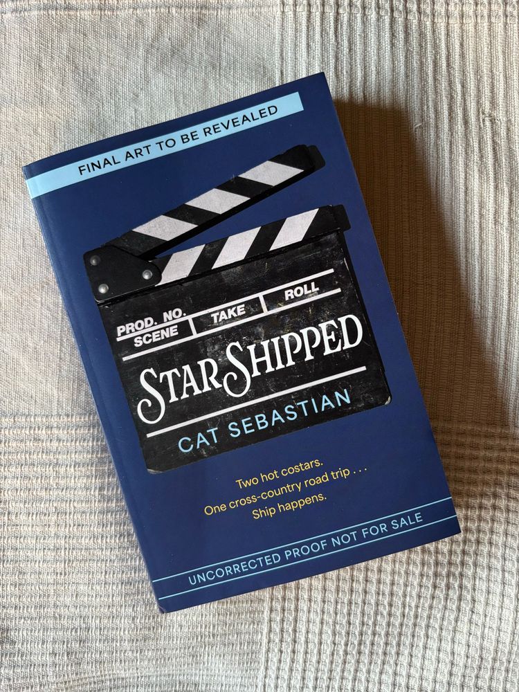 an advance copy of star shipped by cat sebastian on a white blanket. the cover of the book says “final
art to be revealed” and the title and author are pictured on a clapper board. the tagline is “two hot costars. one cross-country road trip. ship happens.”