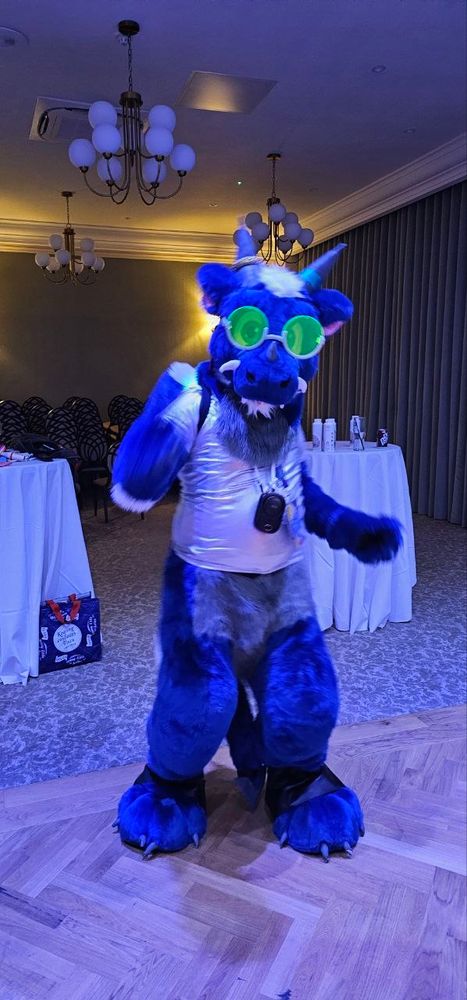 Blue dragon fursuiter Doran dances on a hotel dance floor. He's wearing a silver shirt and florescent green glasses.