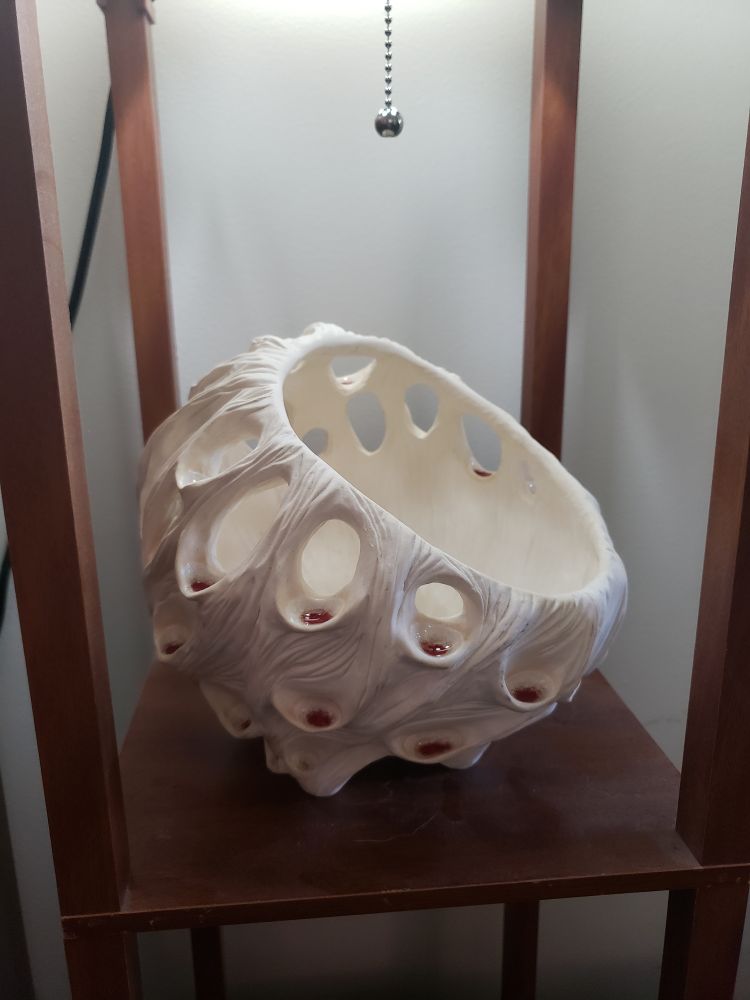 A bone-white, semi-spherical sculpture with a rough textured exterior and smooth interior. Covered in varied sizes of openings that contain little red pools of glass 