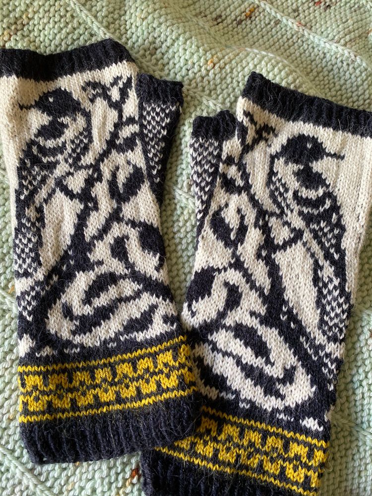 A pair of colorwork fingerless mittens with black and white birds and a yellow cuff laying on top of a mint green shawl. 