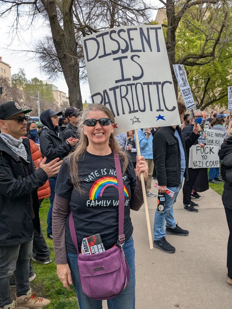 White woman wearing a T-shirt with a rainbow and "Hate is Not a Family Value" on it carrying a sign reading "Dissent is Patriotic" 