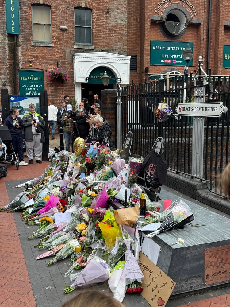 Bunches of flowers cover a bench on a bridge with cut out figures of four men on it. The railing behind has a sign which reads Black Sabbath bridge