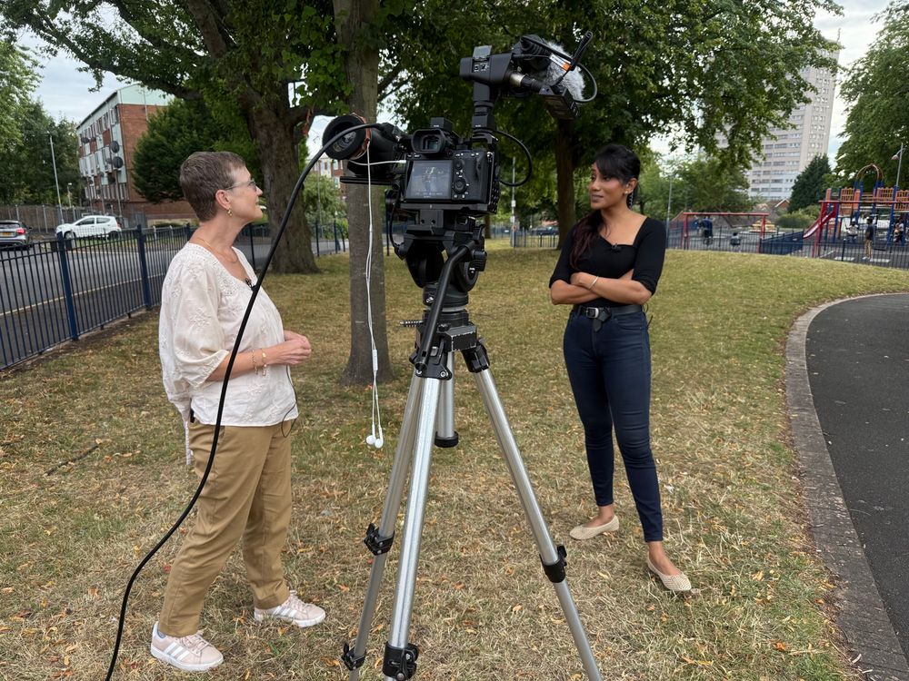 A woman with short hair and glasses is standing facing a woman with long dark hair who is facing away from the camera. There is camera equipment visible. There is a railing, a road, trees and a block of flats beyond 