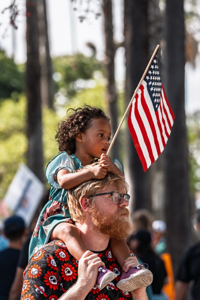 Anaheim, CA – June 14, 2025: A young girl sits atop her guardian’s shoulders, waving an American flag during the No Kings protest. The peaceful demonstration drew families, children, and community members of all ages, creating an open and inclusive space for civic expression.