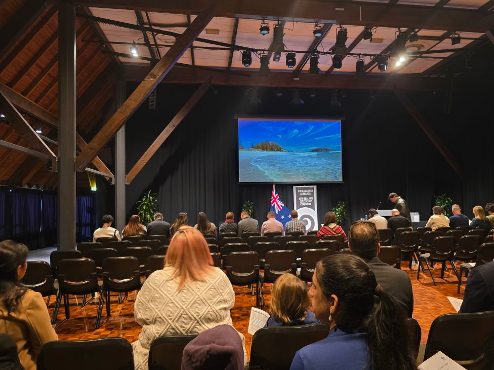 Image shows a hall with people sitting in chairs waiting. There is a screen above the stage showing an image of Mt Maunganui.