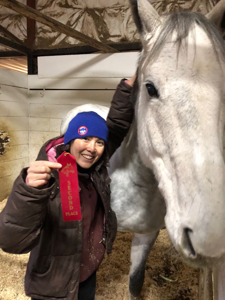 A happy middle aged Asian woman holding a 2nd place ribbon won by a steel grey horse (Woody) who stands beside her.