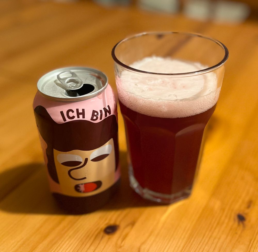 A can of Ich Bin Raspberry beer placed on a wooden table next to a glass filled with the pinkish-red beer.