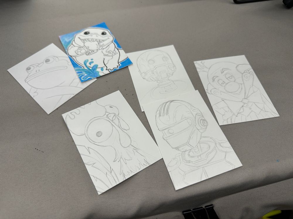 Pencil sketches of cartoon characters on paper, including a frog, chicken, robota, and a blue-colored shark drawing, are displayed on a gray surface.