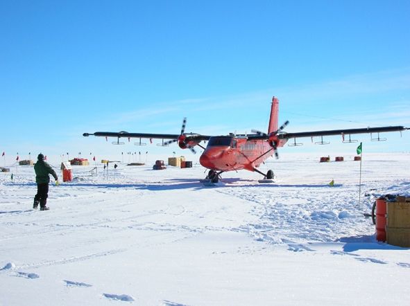 A red aircraft sitting on white snow surrounded by boxes. The sky is blue and it is sunny. A man in dark clothing is standing in front of the plane. Radar antennas can be seen attached below the wings. Photo credit: British Antarctic Survey (BAS)