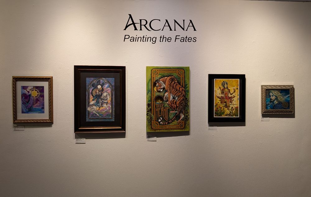 Photo of a gallery wall with Arcana: Painting the Fates above five paintings, from left to right a watercolor painting of 'the Star' by Naomi Van Doren, a knight by Matthew Spencer, a tiger representing the Hermit by Jaime Thompson, the Magician by Sean Andrew Murray, and a Celtic knight by Jody Lee