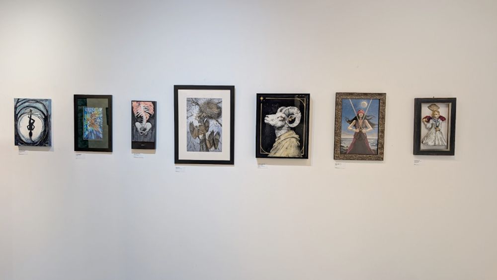 View of a gallery wall featuring art by Christy Lynne Seving of a hanging man in an abstract swirling background my art of a 2 of pentacles fox anthro over stained glass @dorywhynot.bsky.social watercolor piece of a spine and hips on fire @windfalcon.bsky.social of two bald eagles falling from a next that is falling from it's weight breaking the tree Sharlene Loomis The Hermit ram headed anthro looking into a black sky @melissagayyy.bsky.social 2 of swords of a woman blindfolded with crossed swords  and Penney Bidwell Temperance sculpture of a winged woman in a frame
