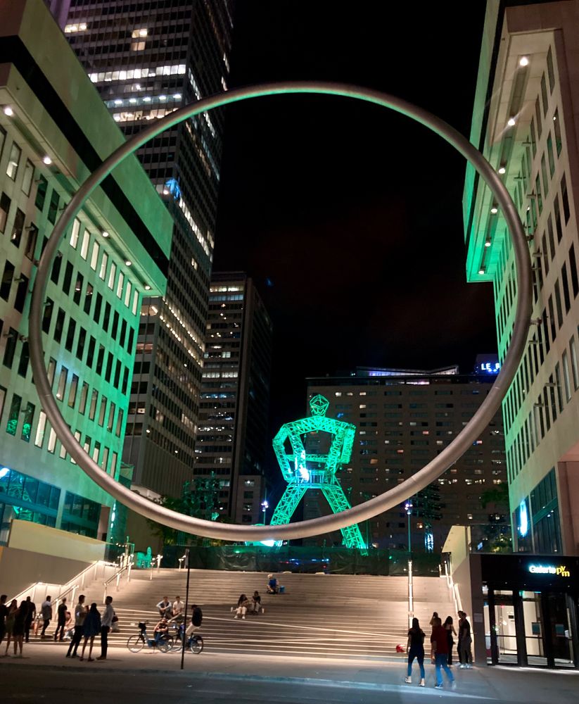 Photo of an art installation in downtown Montreal, of a large metal ring suspended between two buildings, with a large glowing green humanoid figure in the background. There is a large wide staircase at the base between the two buildings and The Ring.