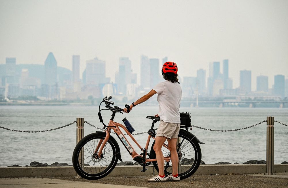 Photo shows a bicyclist looking across the water to the skyline of the city of Montreal, which is barely visible through the hazy smoke from western wildfires.