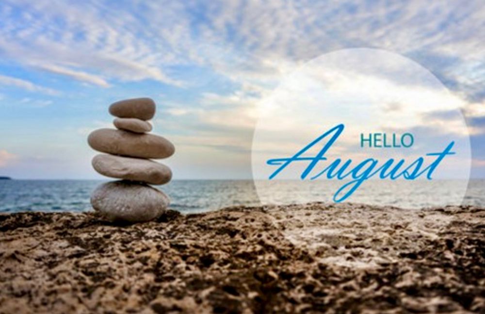Image shows a rocky beach, with 5 stacked rocks forming a tiny tower. The seaside is in the background, and a blue sky with light clouds. There is a text overlay saying “Hello August”