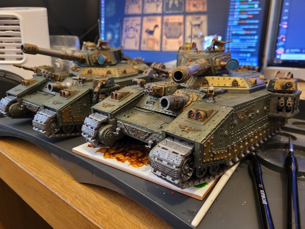 A pair of Warhammer 40,000 Astra Militarum Baneblade models, sat on a desk. Their paint job is a mixture of dull green, with brass and gunmetal paneling, weapon casings and tracks with occasional red and blue details.