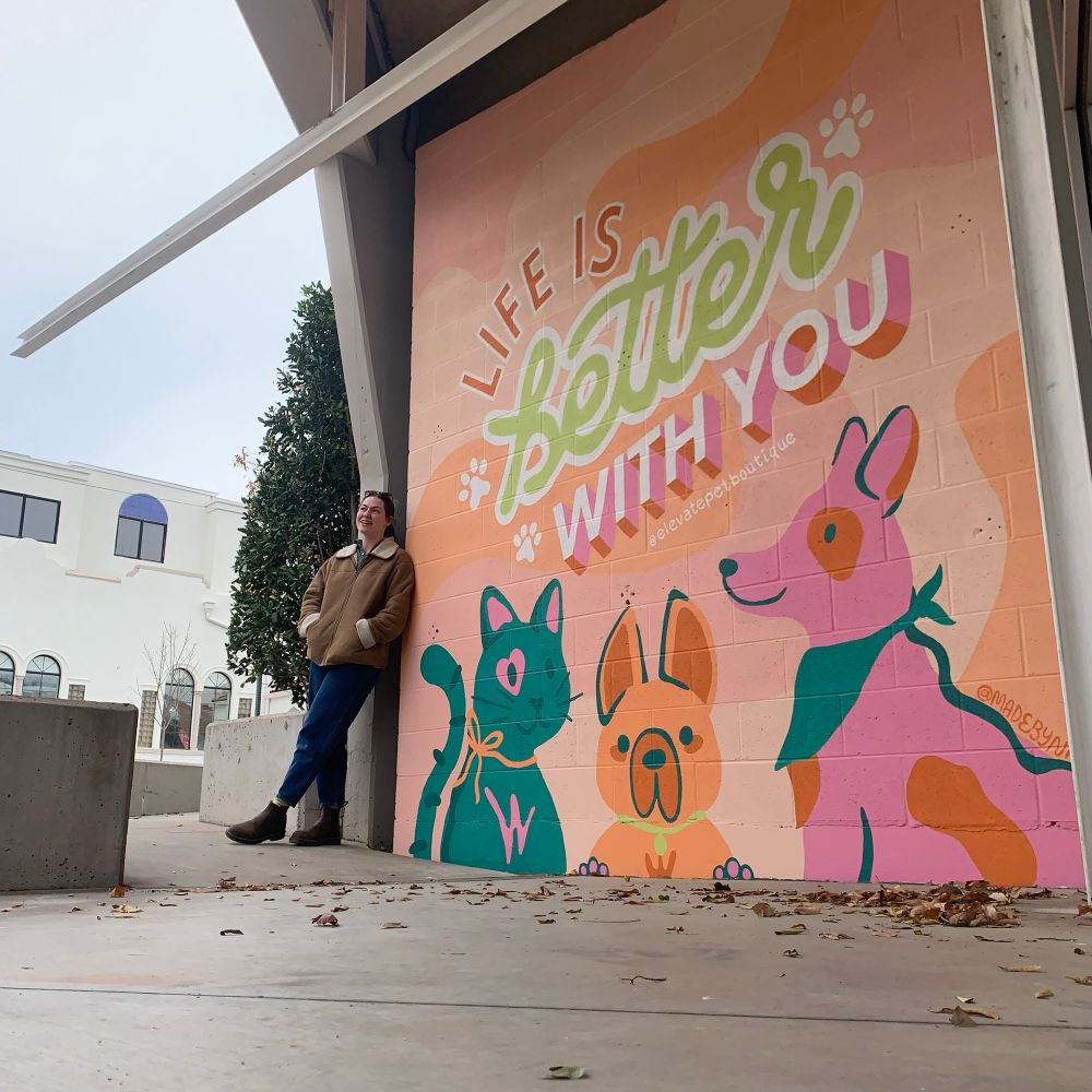Nat stands next to a mural she painted that says “life is better with you.” There are three colorful pets beneath the quote: a teal cat with pink markings (including a pink heart around one eye) and an orange bow, an orange french bull dog (I think) with a green collar, and a pink lab with orange markings and a teal bandana. 