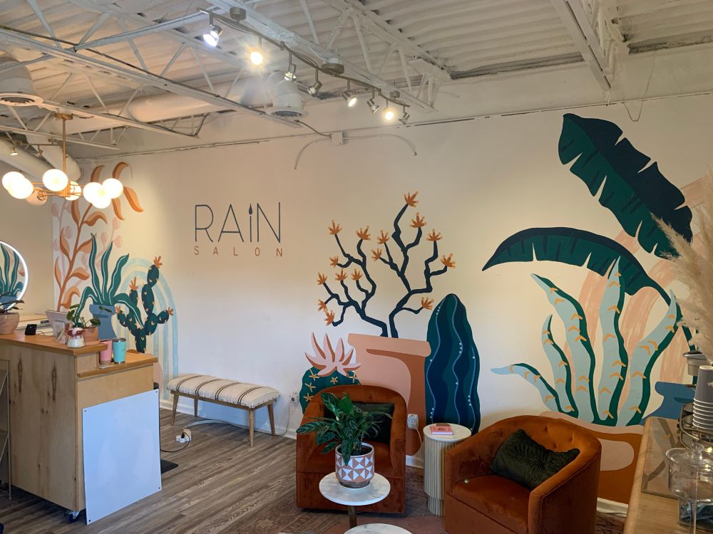 A mural full of potted plants, including palms, snake plants, succulents, and cacti. Dark teal, dark blue, light blue, terra cotta, pink, orange, and yellow are the main colors. The mural also includes the logo for Rain Salon. You can see the cute salon sitting area and reception desk as well.
