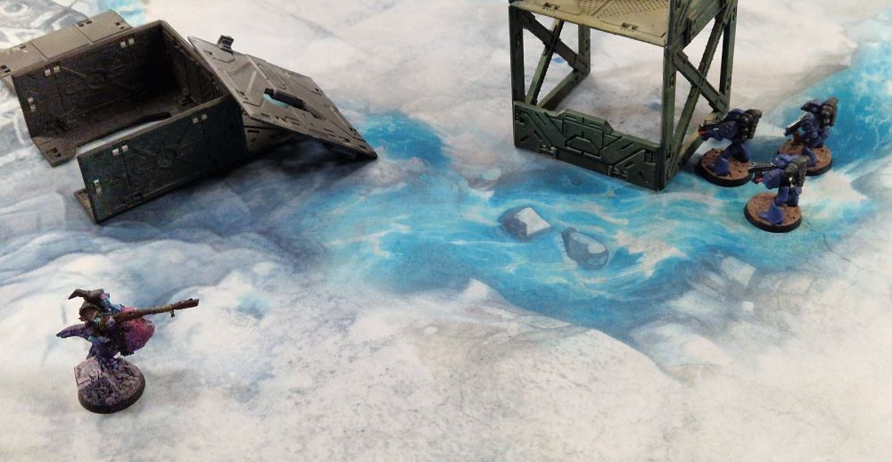 Three space marines face up to a goblin on some sort of hopper, in a snowy wasteland littered with the remains of some prefab buildings.