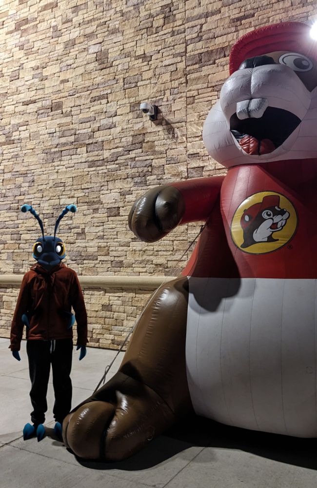a photograph of me standing beside a Buc-ee's inflatable, ominously lit. I've digitally imposed a 3D model of my ant VRchat avatar over myself and lit it appropriately
