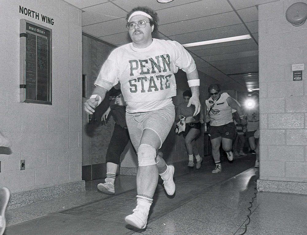 black and white image of a large man in workout gear (gym shorts, sweatbands, mesh Penn State shirt) running down the hall