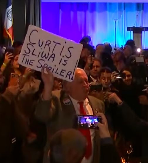 an older white gentleman in a ill-fitting suit holding up a hand-written sign that reads "CURTIS SLIWA IS THE SPOILER!"