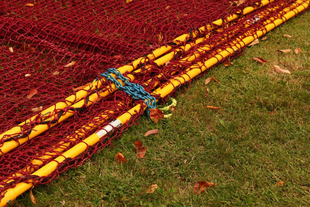 A photo of some red netting lying on green grass. The net is held together by some yellow pipes that cross the frame diagonally, from bottom left to top right. This makes the photo roughly half-net half-grass. In the center, a blue chain connects the pipe to a bright green anchor. I feel like I've posted this one here already but oh well lol