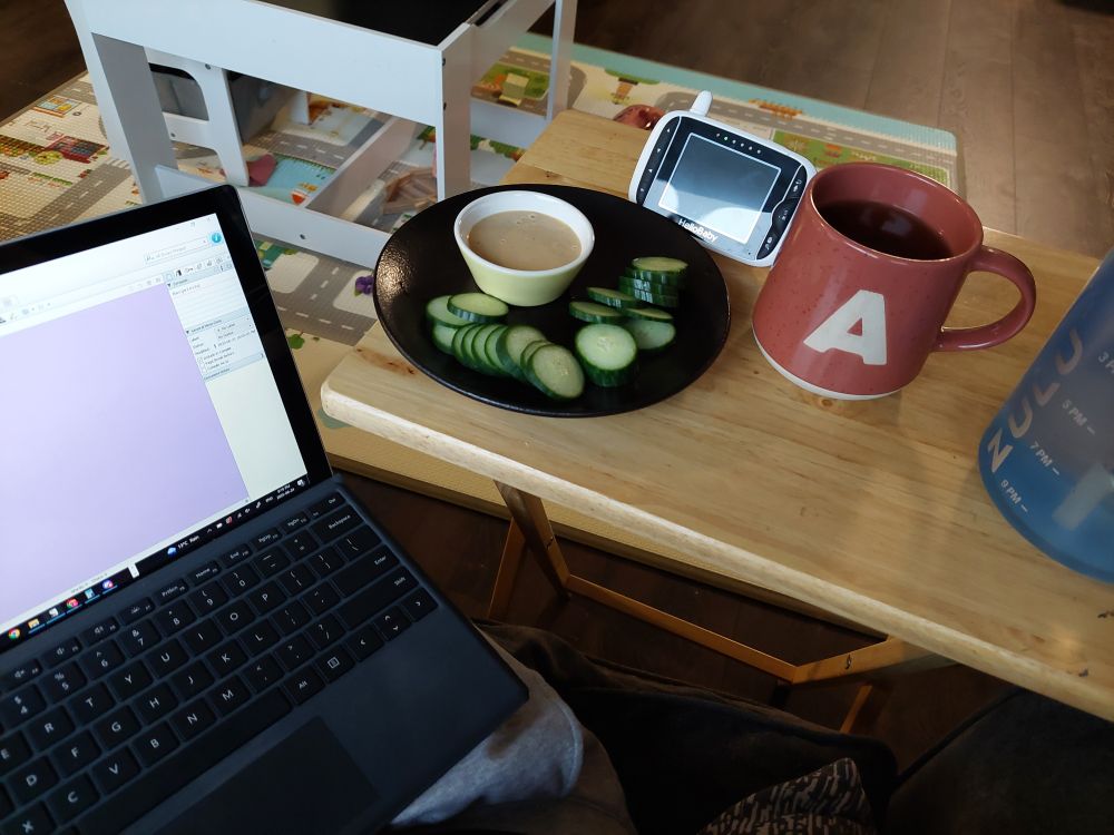 A picture of a Laptop open to Scrivener, cucumber with dip, baby monitor, cup of tea, and a water bottle.