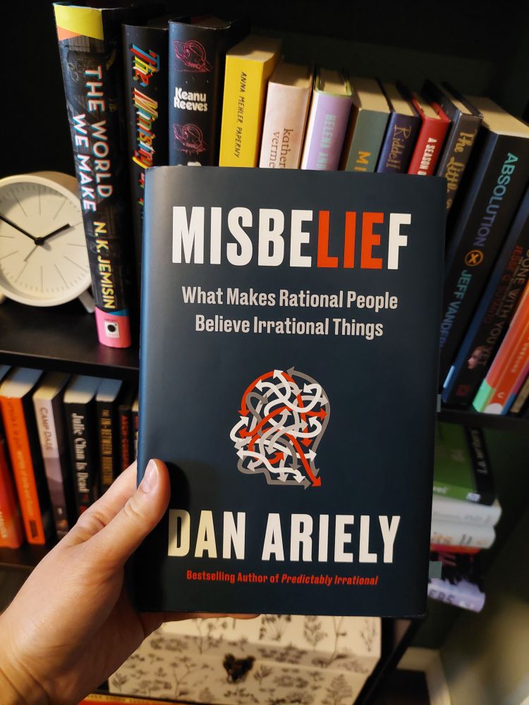Book held in front of bookshelf, "Misbelief" by Dan Ariely