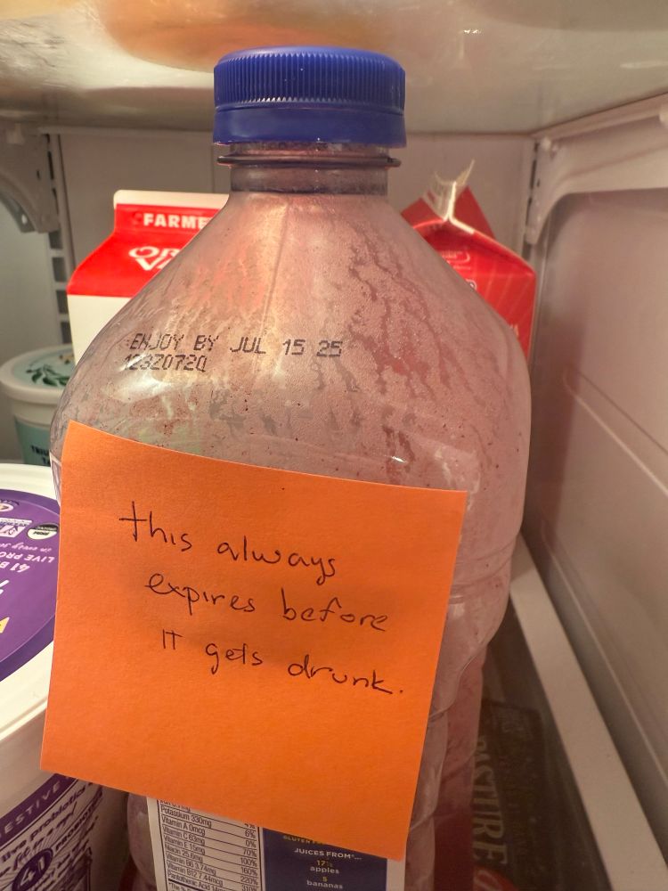 A large expired bottle of juice in a fridge with a post it reading “this always expires before it gets drunk”