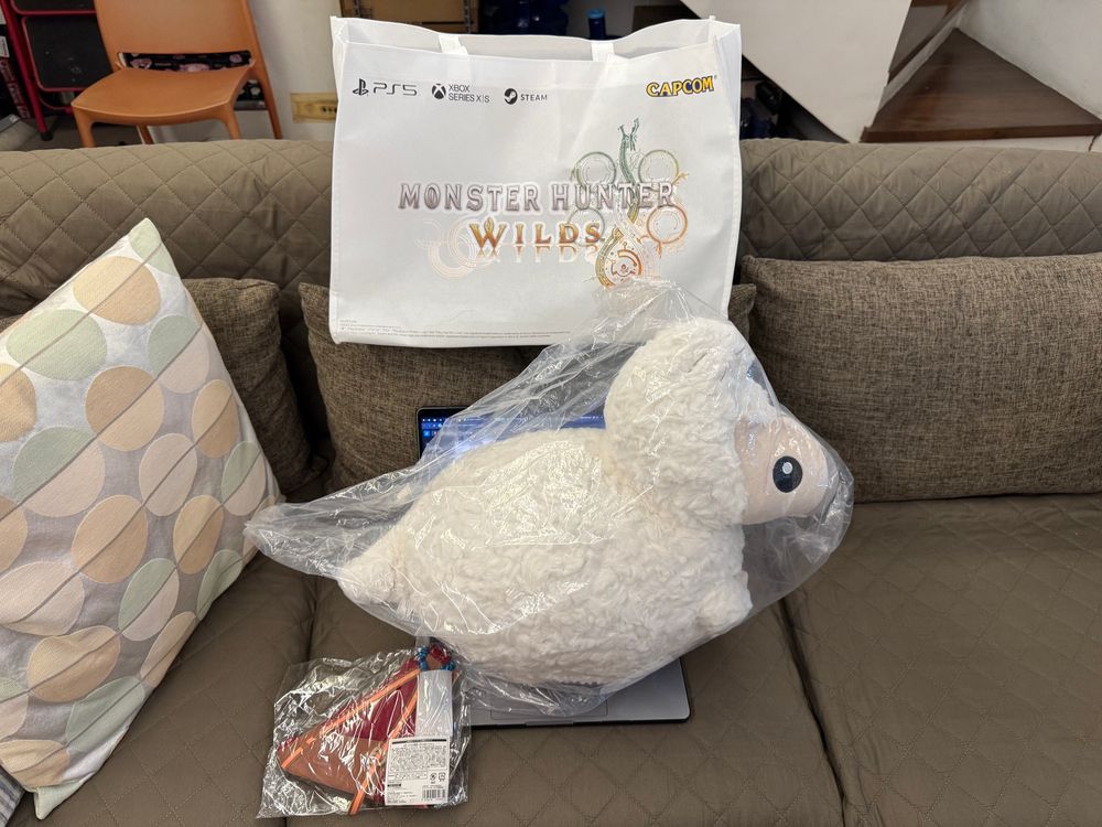 A huge plushie of a Baby Seikret from Monster Hunter Wilds in its shipping plastic placed on top of an opened MacBook Pro 14"