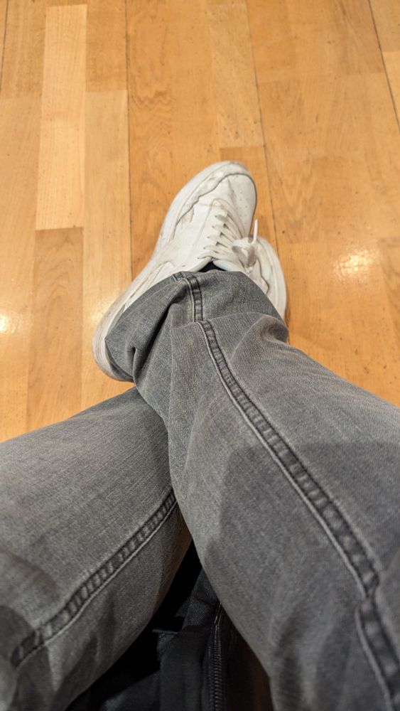 Aufsicht auf überkreuzte Füse mit grauer Jeans und abgetragenen Reebok Sneaker weiß. Eichenfarbener Wartezimmerfußboden im Hintergrund. 