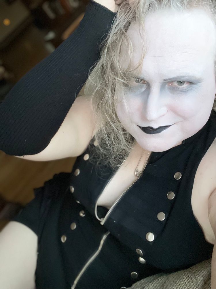 Bell in black and white makeup and an all black outfit. She smirks as she plays with her hair. 