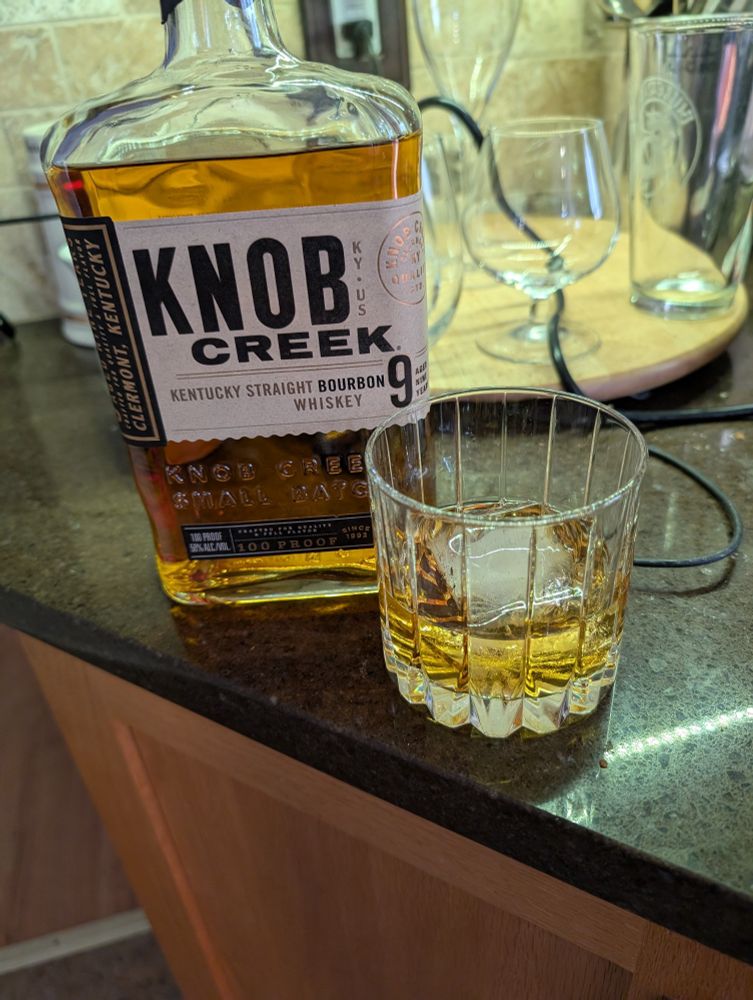 A fifth sized (750 ml), of Knob Creek bourbon next to a 2 ounce pour of said bourbon over a large ice cube.